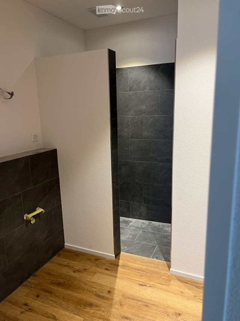 Modern bathroom with black tiled walls and wooden flooring, featuring a sleek black vanity and a large shower area.