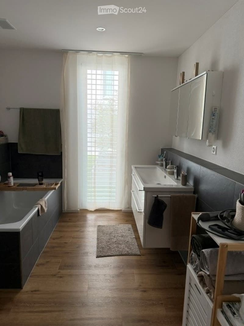 Modern bathroom with a bathtub, sink, mirror, and wooden floor. Features a curtained window, a small rug, and a towel rack with towels.