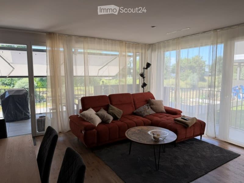 Bright living room with large windows and sliding glass doors leading to a balcony. Features a red sectional sofa with multiple throw pillows, a round coffee table, and a modern floor lamp.