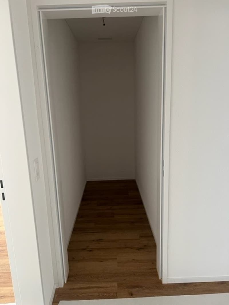 Empty hallway with white walls and wooden flooring, leading to another room.