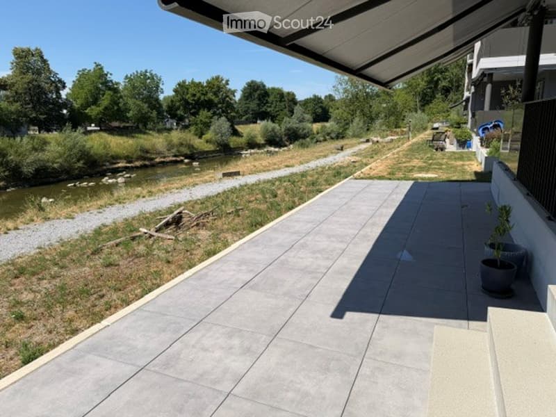 A modern, covered balcony with concrete tiles, overlooking a serene river and green landscape.