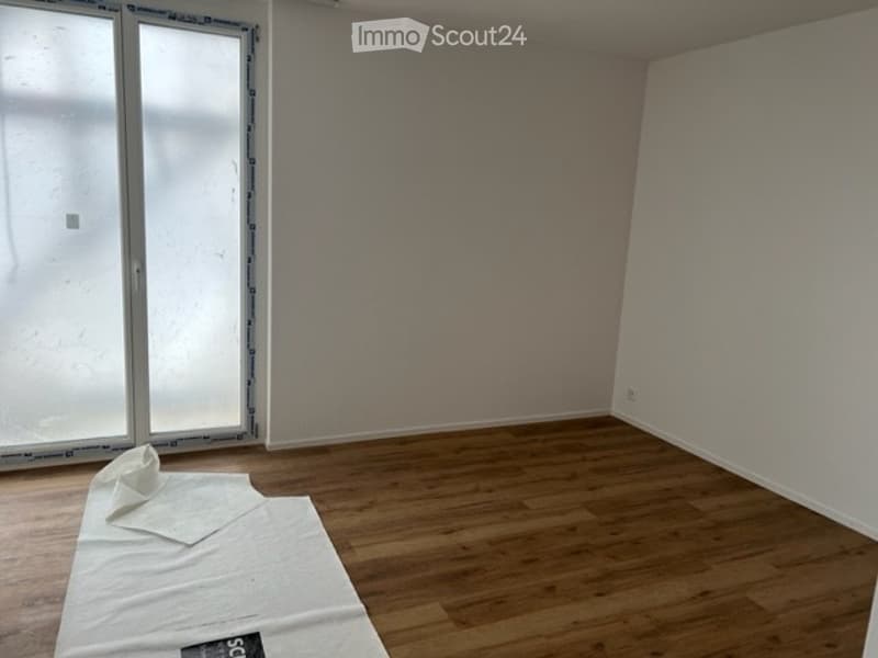 Empty room with wooden flooring and large windows, showing a partially painted wall and a white drop cloth on the floor.