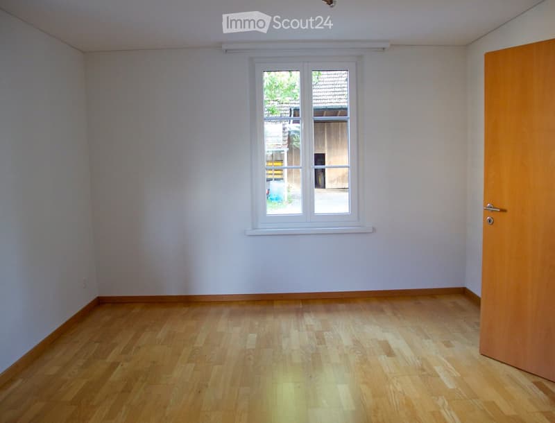 Empty room with light wooden flooring and white walls, featuring a window and a door.