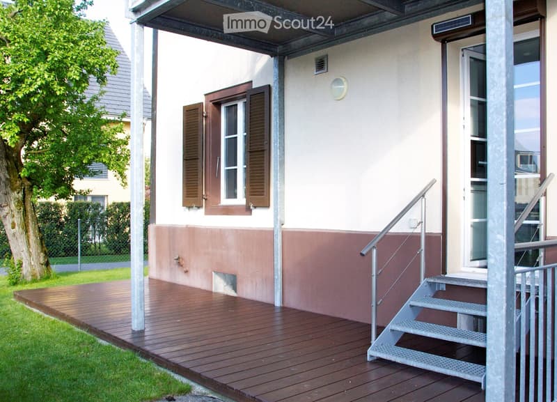 Wooden deck with metal stairs leading up to a house entrance, featuring a window with shutters and a small light fixture above the door.