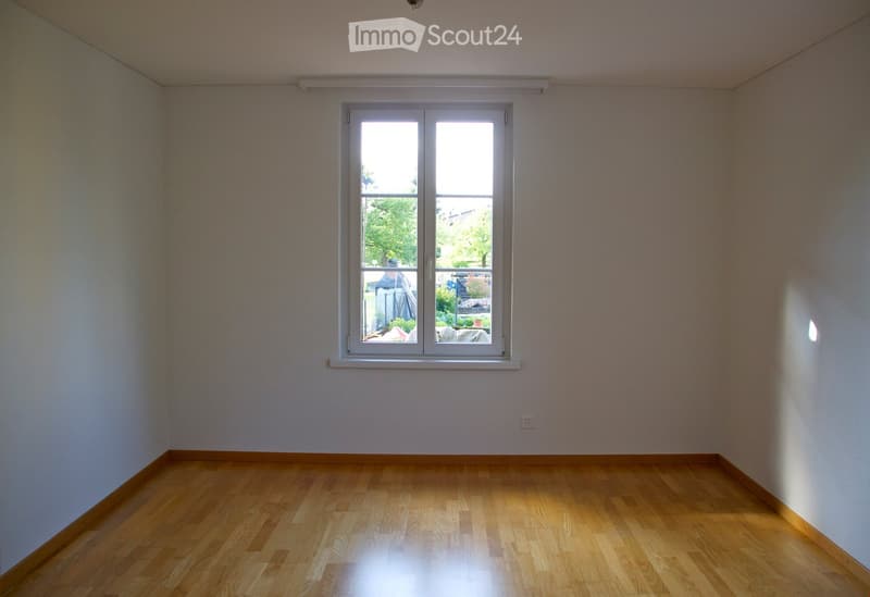 Empty room with wooden flooring and white walls, featuring a large window with white frame and a view of the outside garden.