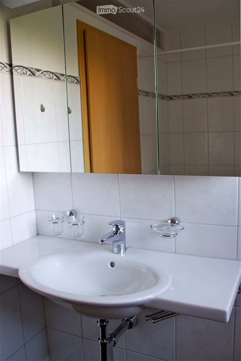 Modern bathroom with a white sink, chrome faucet, and mirror above. Two small cups are placed on the sink counter, and a glass shelf holds additional cups.
