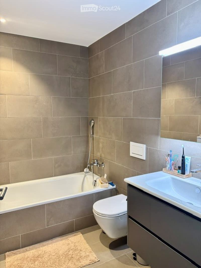 Modern bathroom with beige tiles, a bathtub, toilet, sink, and a mirror above the sink. A small shelf holds toiletries.