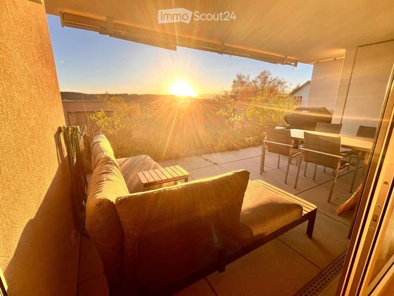 Balcony with a sofa, chairs, and a table, overlooking a scenic sunset view with greenery and a barbecue in the background.