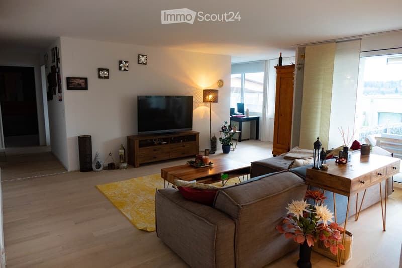 Modern living room with wooden flooring, large windows, a grey sofa, coffee table, TV, and decorative items including vases and lamps.