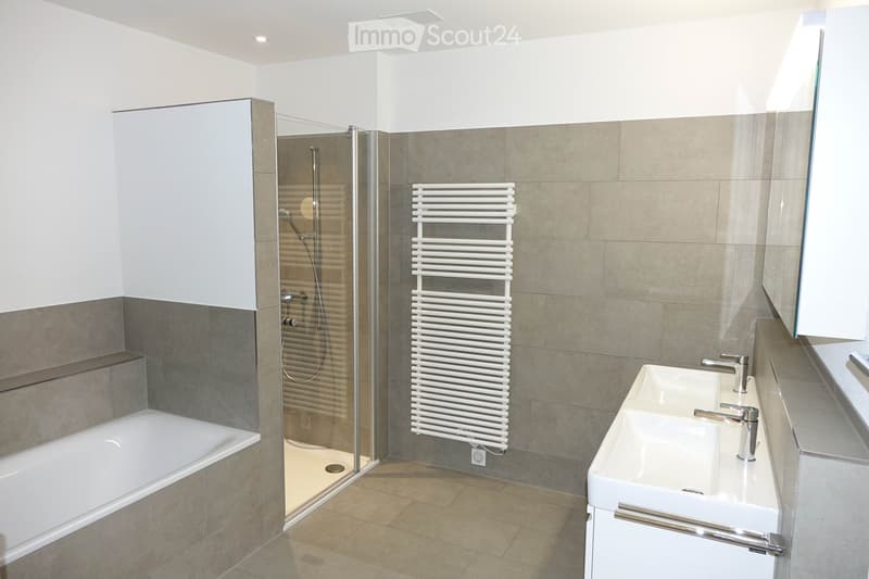Modern bathroom with a bathtub, sink, glass-enclosed shower, and a white radiator. The walls are tiled in gray and beige, and the floor is tiled in beige.