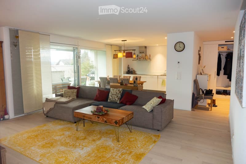 Bright living room with grey sectional sofa, wooden coffee table, dining area with chairs, modern kitchen, and a view through large windows.