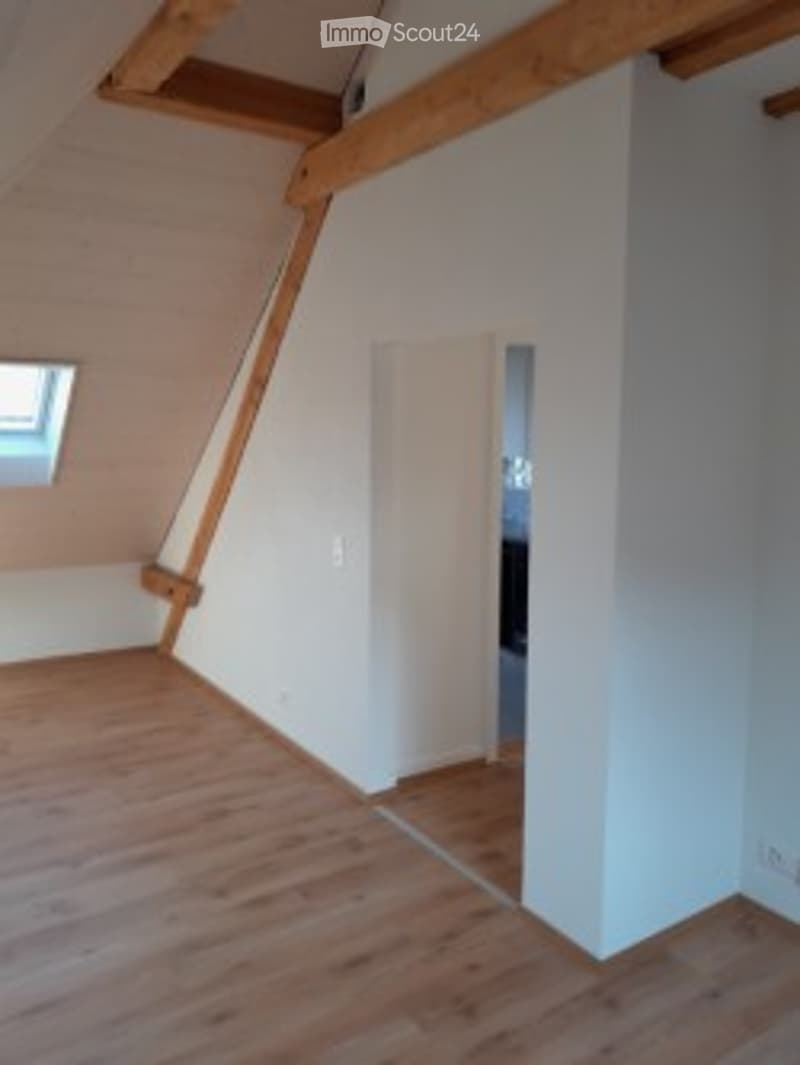 Bright, empty attic room with wooden floors, white walls, and exposed wooden beams. A small window and a white closet door are visible.
