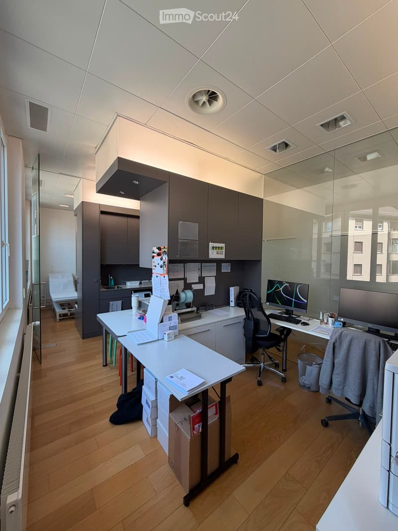Modern office space with wooden flooring, white tables, chairs, laptop, computer screen, keyboard, cabinets, and a small sink area.