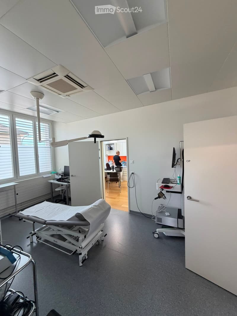 A modern medical examination room with an exam table, chairs, a screen, and a lamp. The room has large windows and a door leading to an adjacent office area.