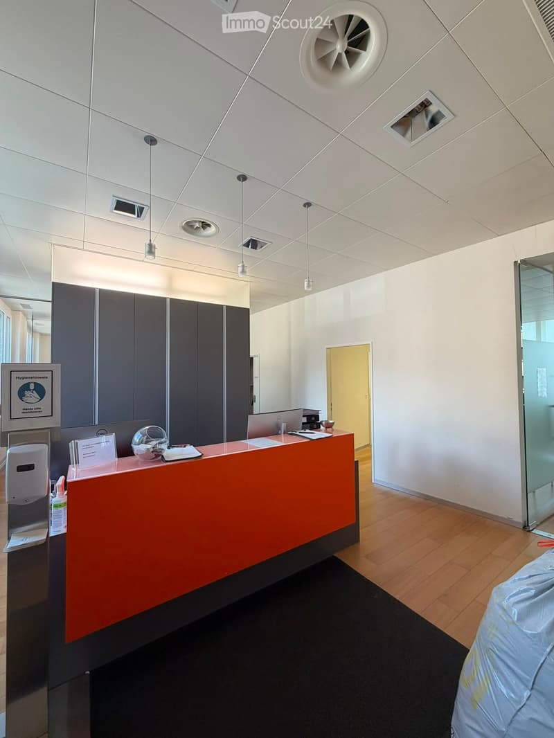 Modern reception area with a bright orange desk, laptop, hand sanitizer, and a sign on the wall. The ceiling features geometric patterns and recessed lighting.