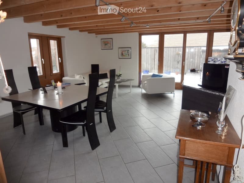 Open-plan living and dining area with exposed wooden beams, large windows, and sliding glass doors leading to a patio. Features a dining table with chairs, a white sofa, a small coffee table, and decorative items on a side table.