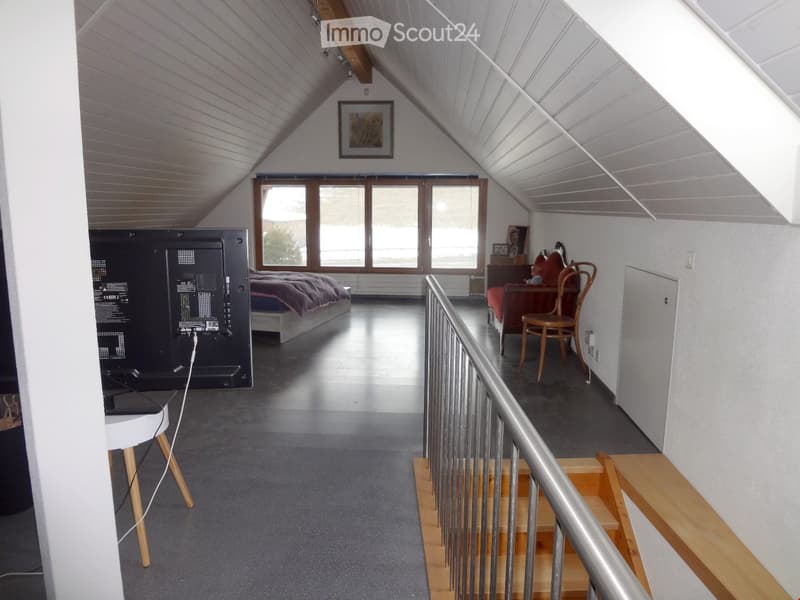Bright attic bedroom with wooden slanted ceilings, large windows, a bed, a chair, a sofa, a small table, and a screen on a stand.