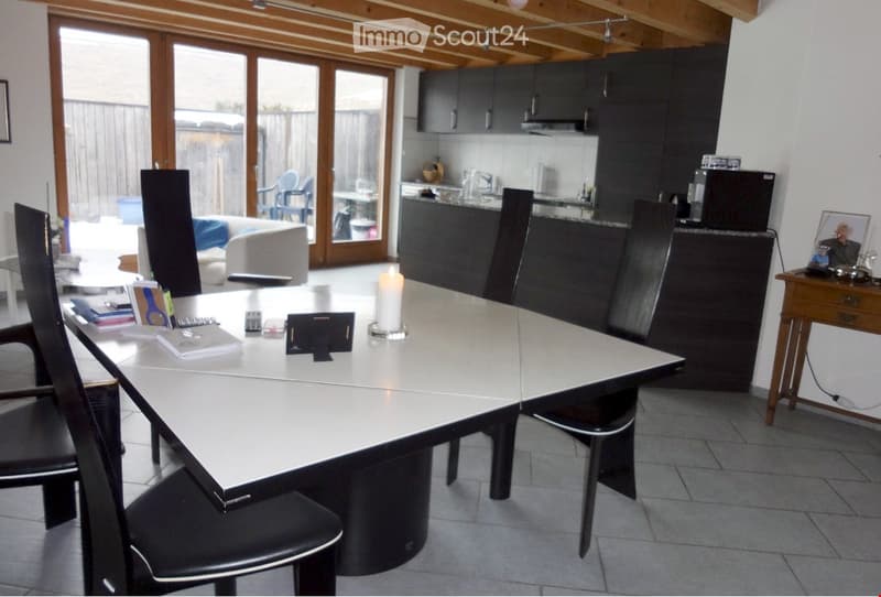 Open-plan room with a large conference table, chairs, a sofa, kitchen area with cabinets and a sink, and a candle on the table.