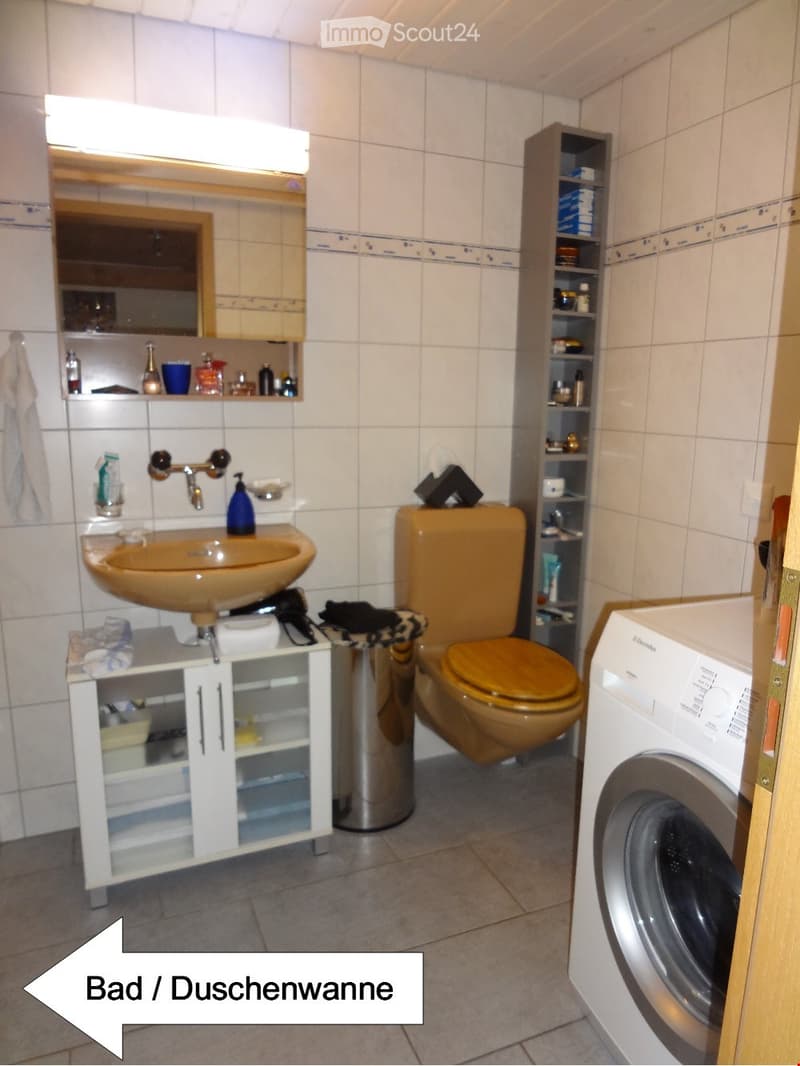 Bathroom with a sink, toilet, mirror, cabinet, washing machine, and shelves. The sink has a modern faucet and a countertop with various toiletries. A small cabinet is placed next to the sink, and a washing machine is visible on the right side.