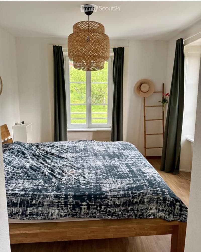 A well-lit bedroom with a wooden bed, patterned duvet, a hanging woven lamp, a wooden chair, a small table with a device, and a hat on a wooden stand near the window.