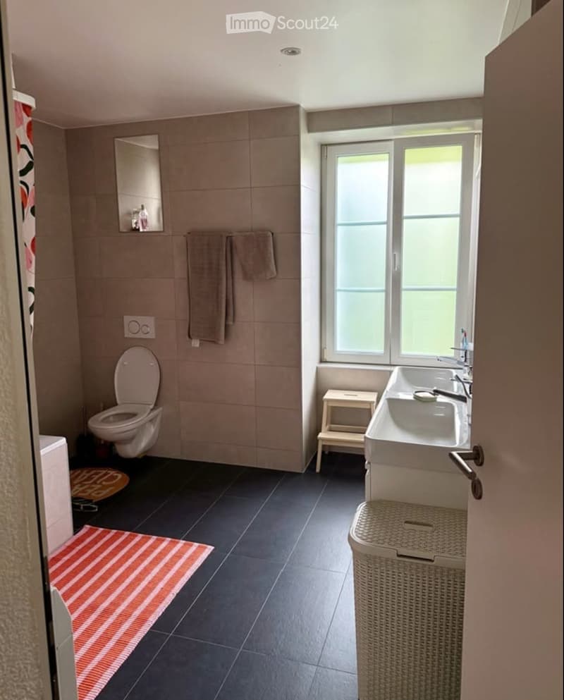 Modern bathroom with beige tiles, a toilet, a double sink, a mirror, towels, a small stool, and a wicker basket. Features a large window with frosted glass and a striped rug on the floor.