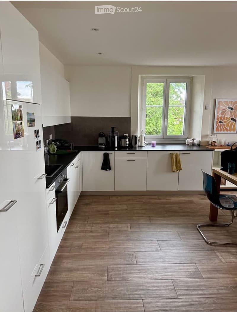 Modern kitchen with white cabinets, black countertops, and wooden flooring. Features include a refrigerator, oven, coffee maker, and a small dining area with a chair and table.