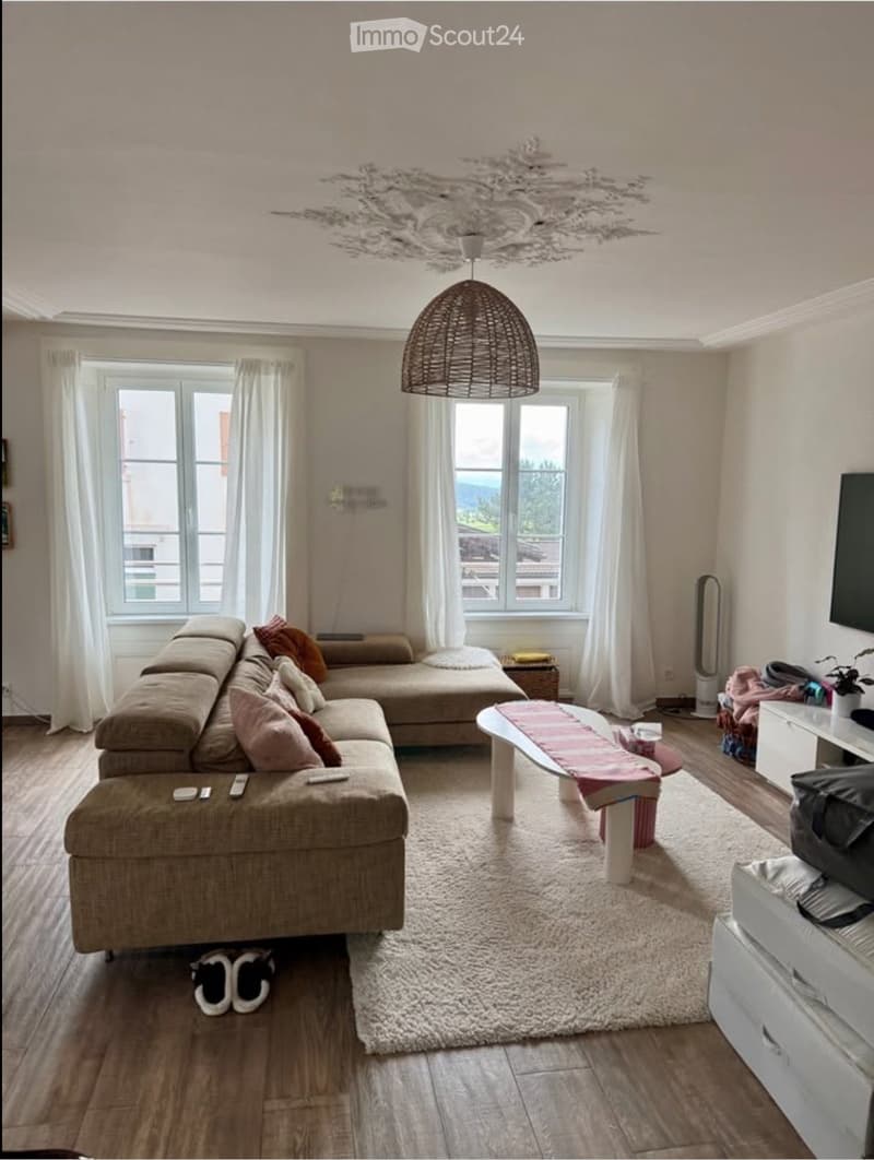Living room with a beige sectional sofa, coffee table, TV, and a hanging woven lamp. The room features large windows with white curtains and a view of the outdoors.