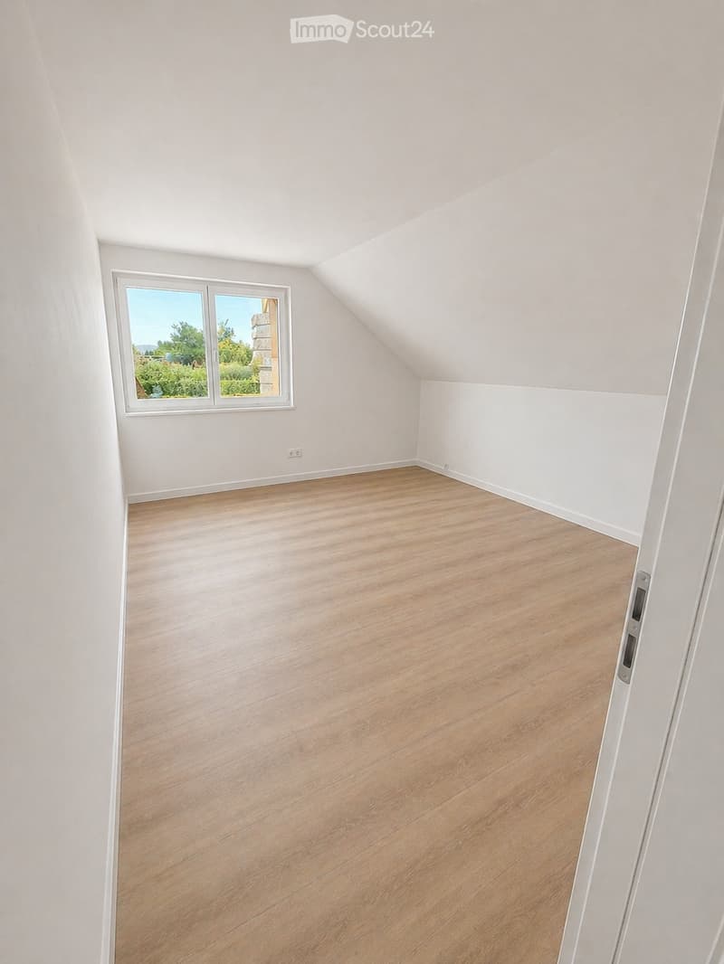 Empty attic room with wooden flooring, white walls, and a large window offering a view of greenery outside.