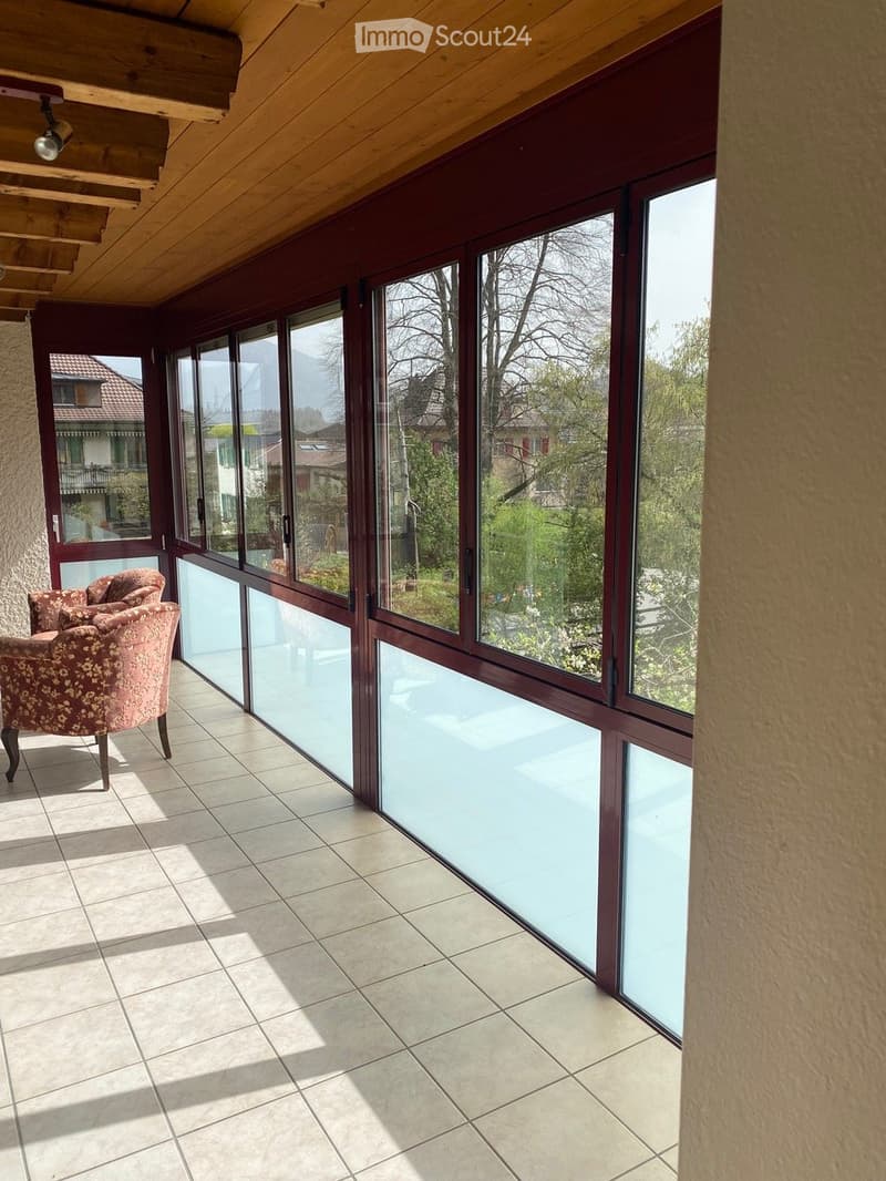 Bright, tiled balcony with large glass windows, wooden ceiling, and a single floral-patterned chair.