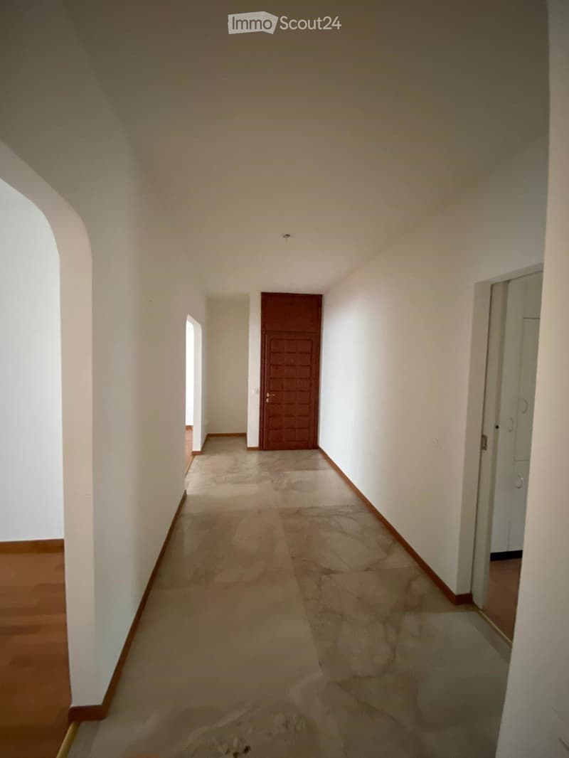 Empty hallway with white walls, wooden baseboards, and a tiled floor. Features a closed wooden door and a partially visible adjacent room.