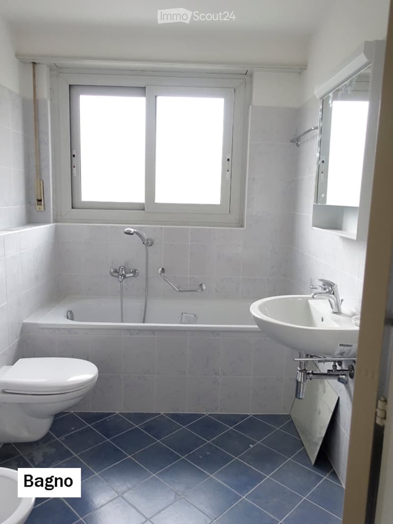 Modern bathroom with white tiles, a bathtub, toilet, sink, and a window providing natural light.