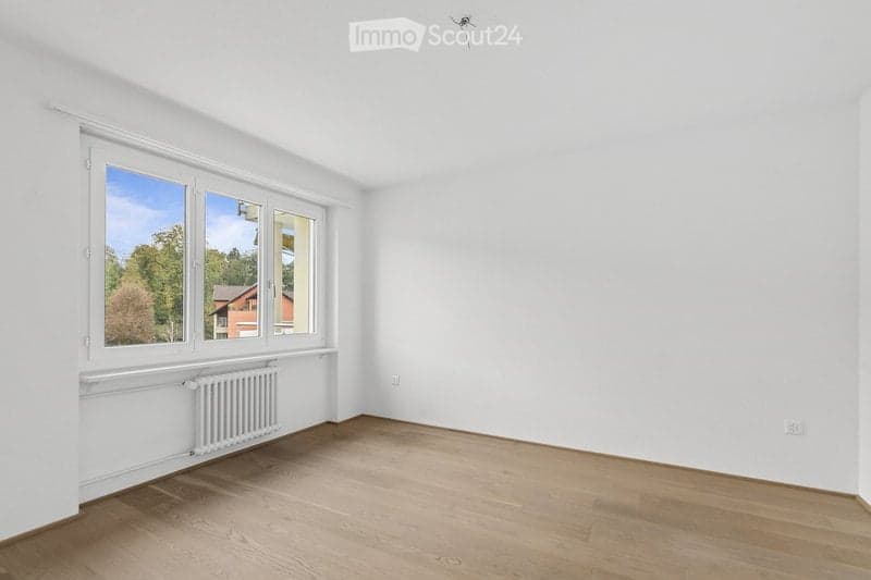 Empty room with wooden flooring, white walls, large windows, radiator, and visible electrical outlets.