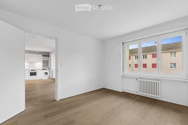 Empty room with light wooden flooring, white walls, and large windows overlooking a street. Adjacent kitchen area visible through an open doorway.