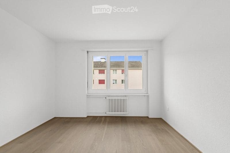 Empty room with white walls, wooden flooring, and a single window with a radiator below it. No furniture or equipment present.