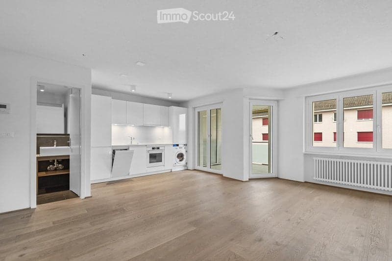Completely empty room with wooden flooring, white walls, and modern white kitchen cabinets visible in the background. No furniture or equipment present.