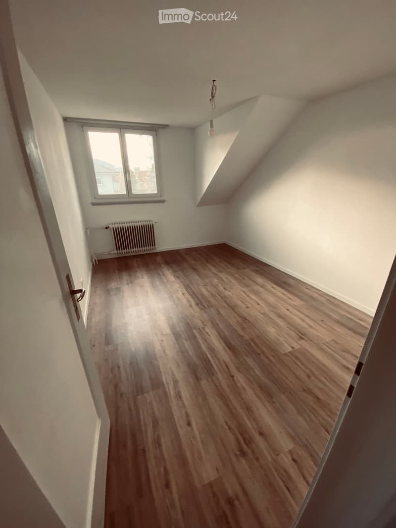 Empty attic room with wooden flooring, white walls, a single window, and a radiator. No furniture or equipment present.