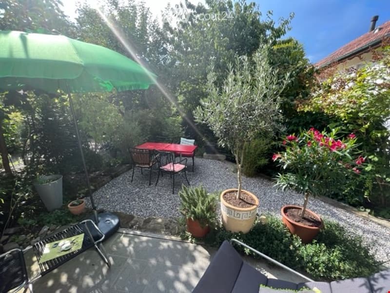 gravel patio with table and chairs, green umbrella, small lounge chair, several potted plants, greenery