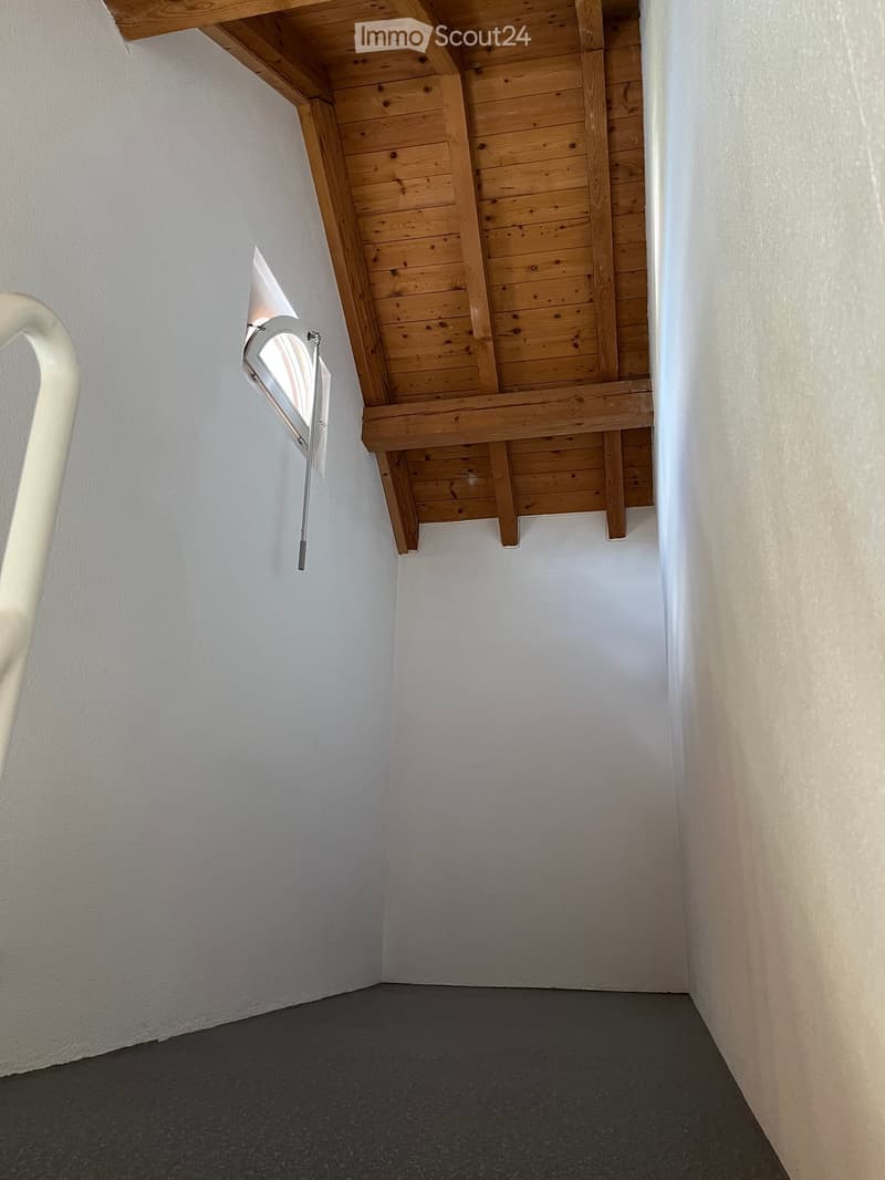 Empty room with wooden slanted ceiling, white walls, and grey carpeted floor. A small window with blinds is visible on one wall.