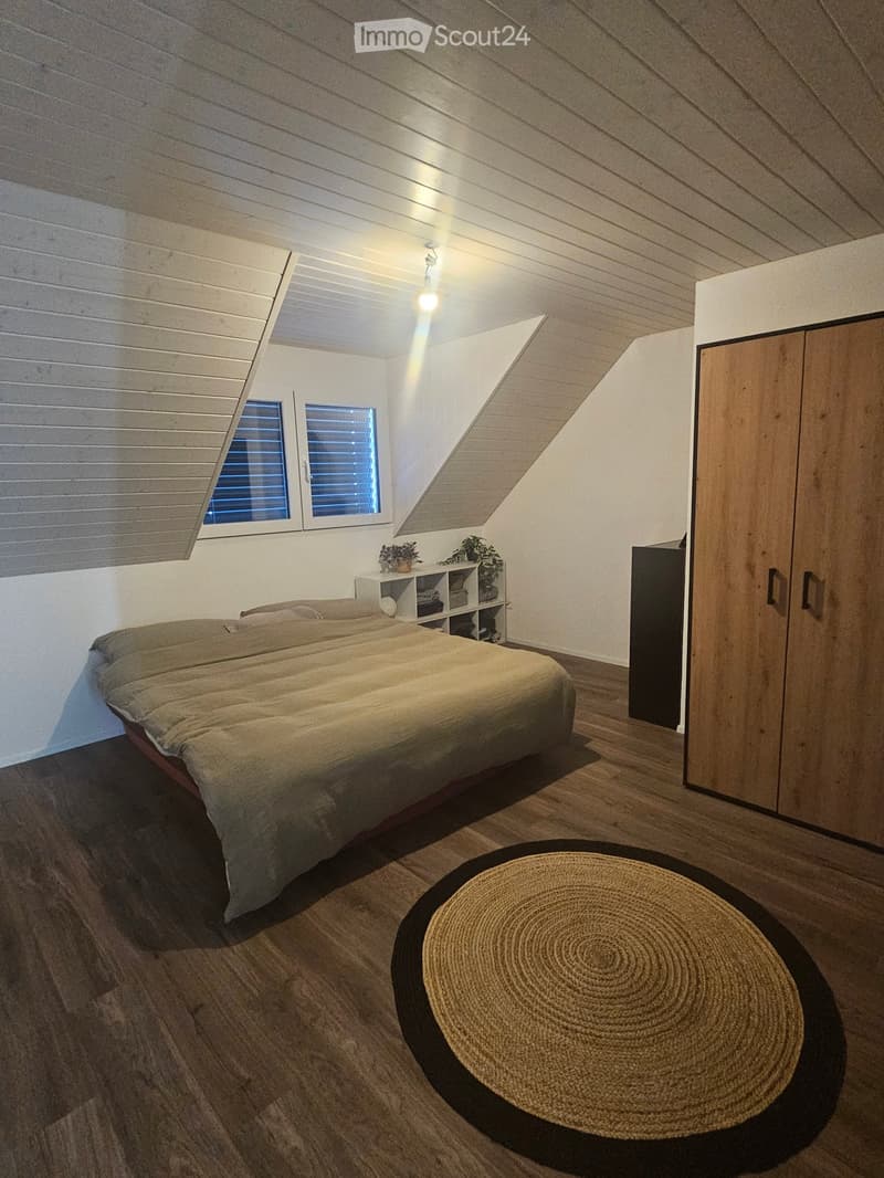 Cozy attic bedroom with wooden slanted ceilings, large windows, a bed, a wardrobe, a nightstand with plants, and a round woven rug on the floor.