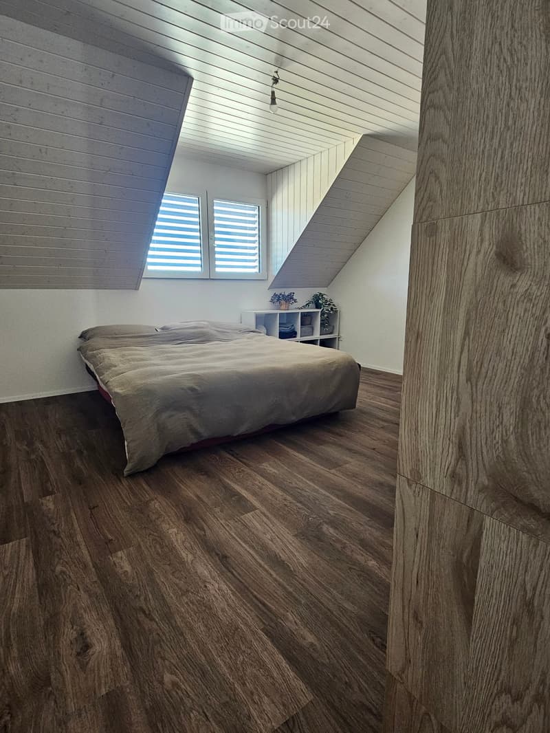 Modern bedroom with wooden flooring, slanted ceiling, and large windows. Features a neatly made bed, a small nightstand with plants, and built-in shelving.