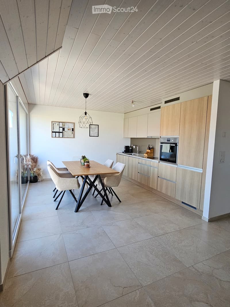 Modern kitchen with light wood cabinets, stainless steel oven, central island, dining table with four chairs, pendant lighting, and decorative wall art.