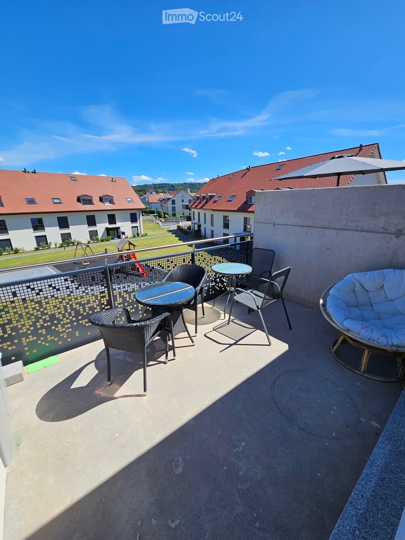 Balcony with a round table, four chairs, a wicker chair, and an umbrella, overlooking a residential area with houses and a playground.