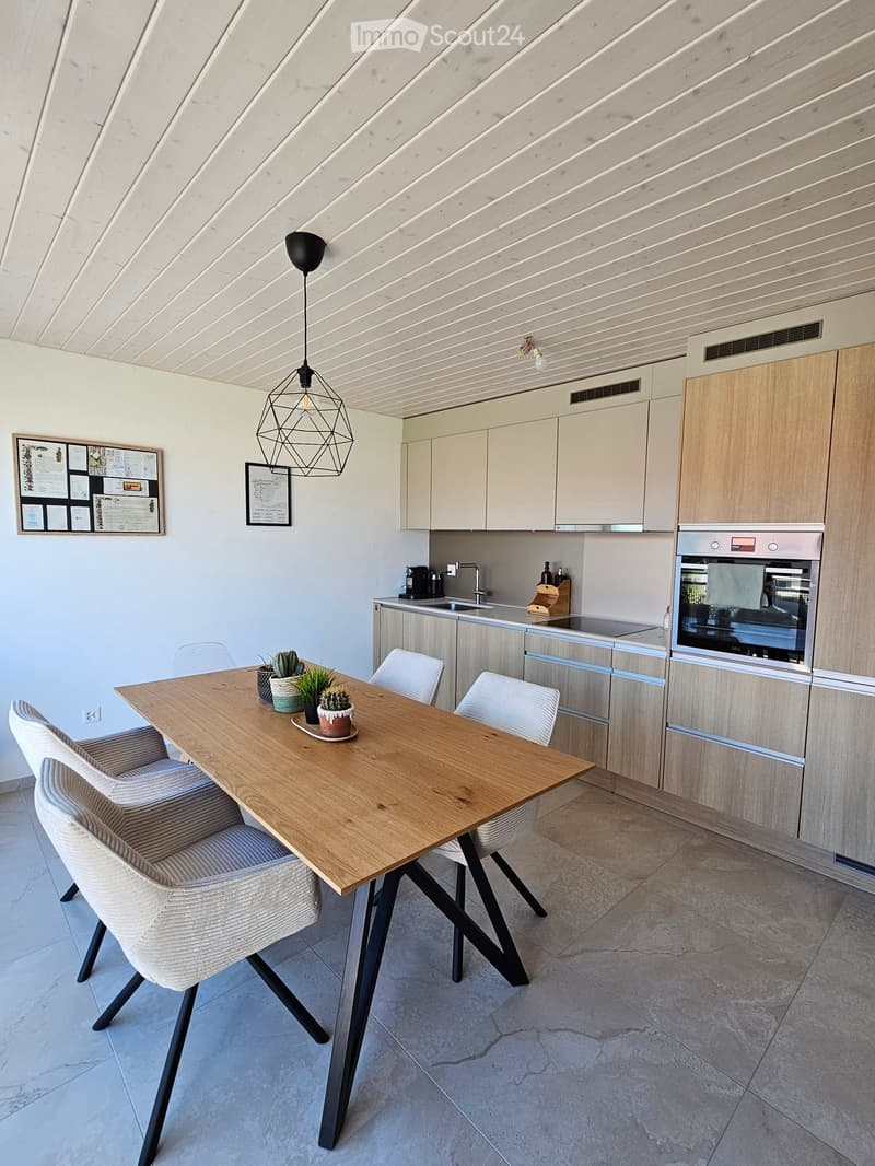 Modern kitchen with light wood cabinets, stainless steel appliances, a wooden dining table with four chairs, and a pendant light hanging above the table. The counter features a sink, faucet, coffee maker, and a small wooden cutting board. There are framed
