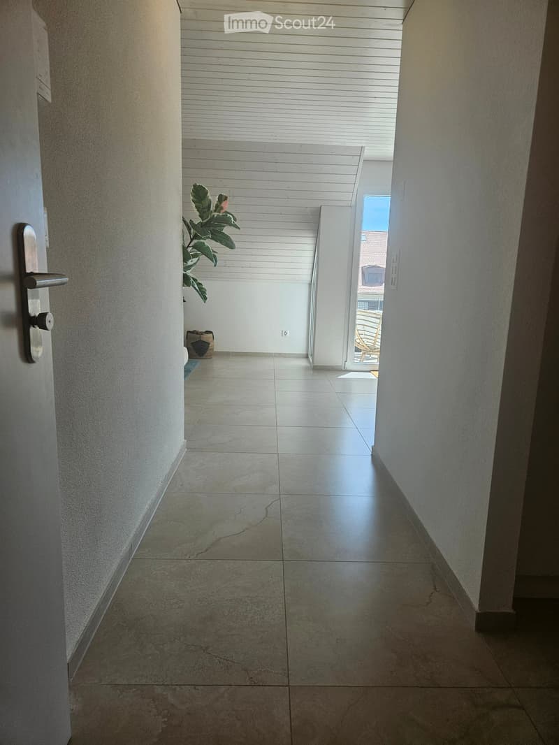 A clean, tiled hallway with a door on the left and an open doorway leading to a balcony area with a plant and outdoor furniture visible.