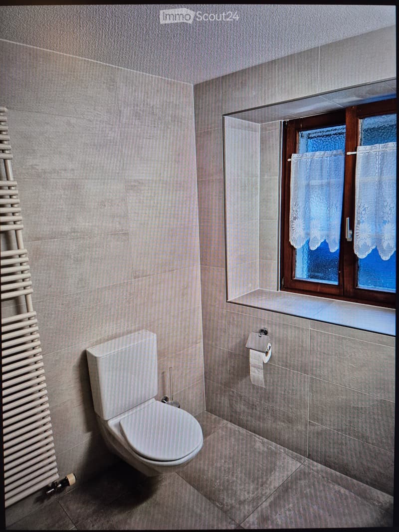 Modern bathroom with grey tiled walls and floor, a toilet, a radiator, and a window with frosted glass and a wooden frame.