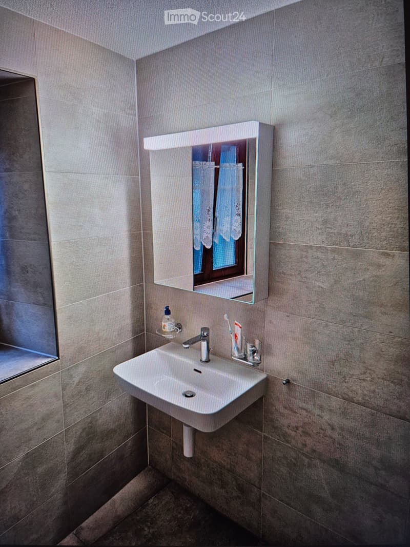 Modern bathroom with a white sink, chrome faucet, mirror with built-in lighting, and toiletries on the sink counter.