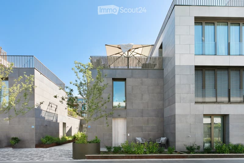 Modern multi-story building with grey concrete facade, large windows, and balconies. Features include a small garden area with potted plants, outdoor seating, and a tree in the foreground.