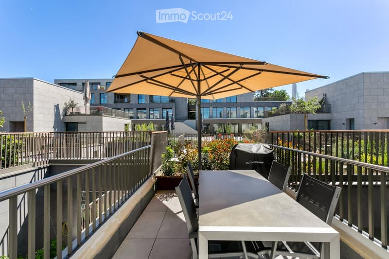 Balcony with a large umbrella, a white table, black chairs, and a covered grill, surrounded by plants and a modern building in the background.