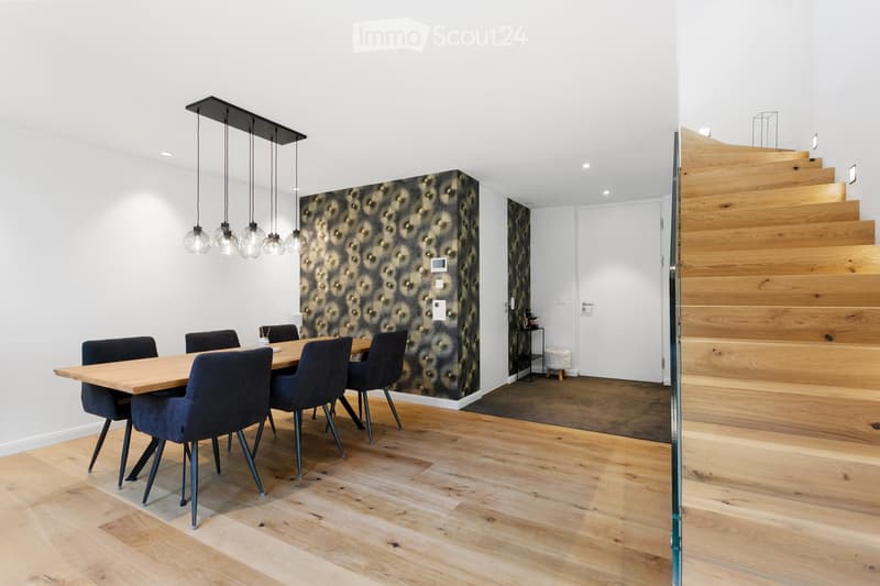 Dining area with a wooden table, dark blue chairs, pendant lights, and a patterned wall. A wooden staircase leads to an upper level.