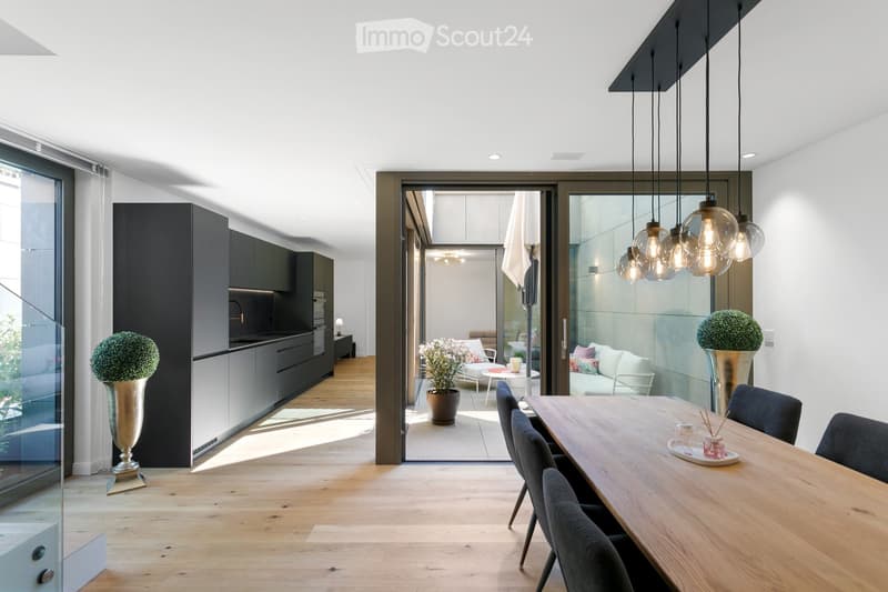 Modern open-plan dining and living area with light wood flooring, large glass doors leading to a patio, pendant lights, a dining table with chairs, a sofa, and decorative vases.